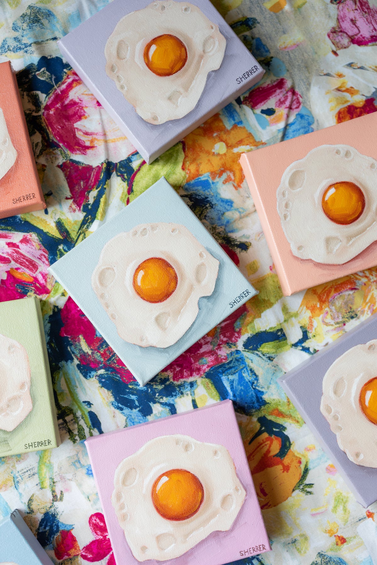Pastel fried-egg paintings arranged diagonally on colorful floral fabric, with a blue painting in the center.