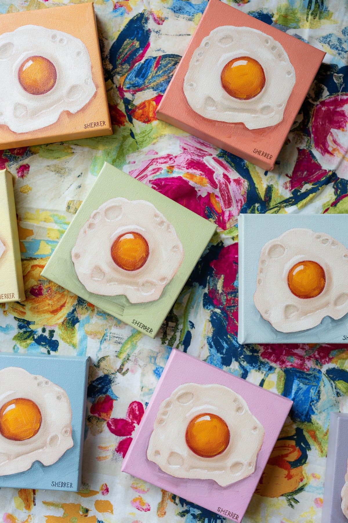 Fried-egg paintings on pastel canvases, with a mint-green painting centered above a pink one on floral fabric.
