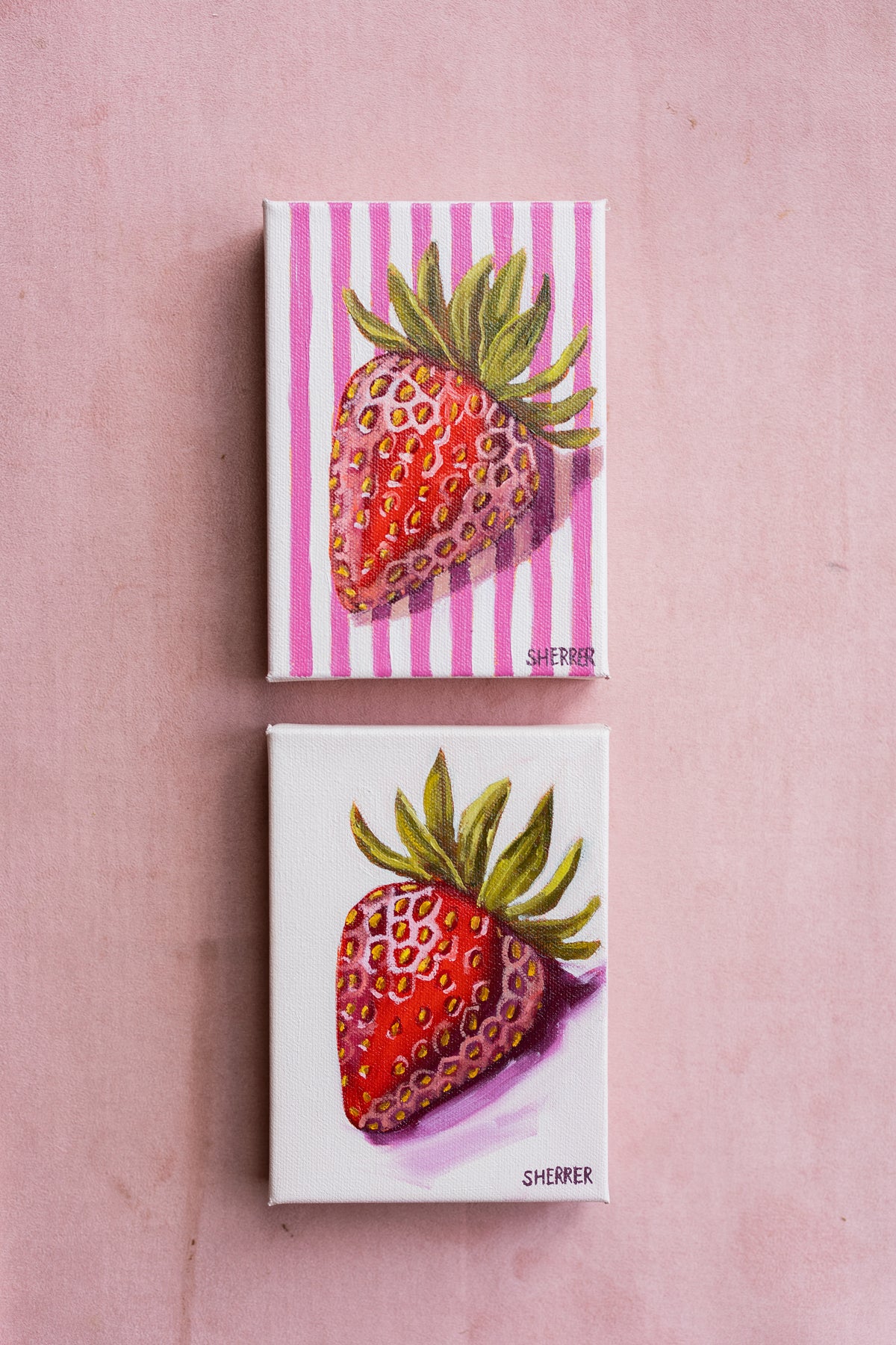 Two small strawberry paintings arranged vertically on a pink surface, one with pink-and-white stripes and one with a pale background.
