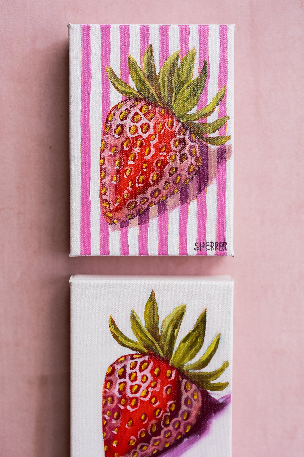 Close-up of two small strawberry paintings on a pink surface, with striped and pale backgrounds.
