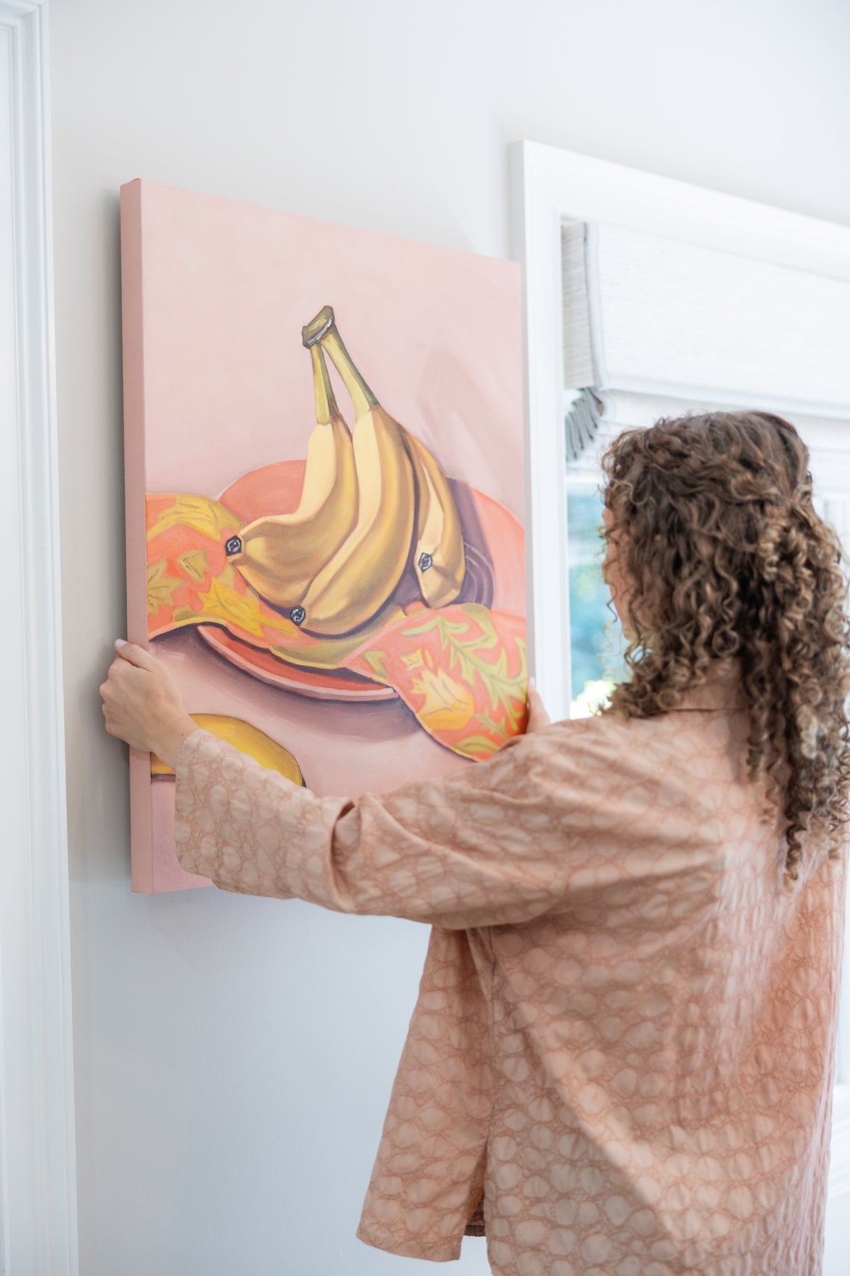 Caroline Sherrer positioning Bananas and Botanicals II, a banana still life on pink, on a wall.