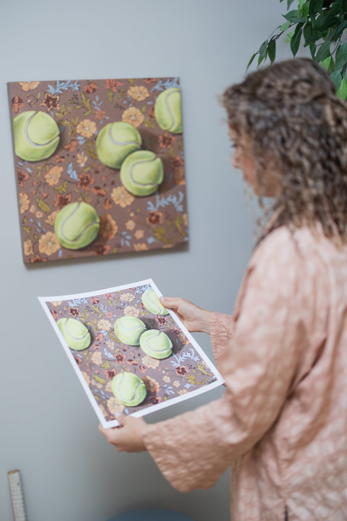 Caroline Sherrer holding a white-bordered Weather for Tennis print beside the matching tennis-ball painting on a floral background.