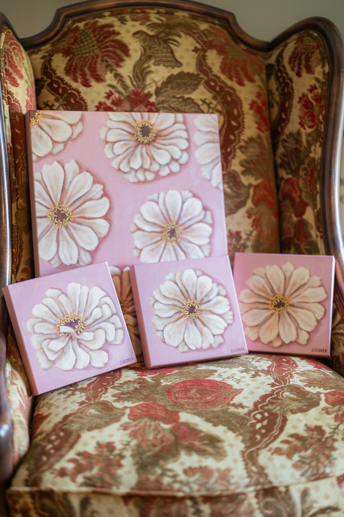 Angled view of three small white-zinnia paintings and a larger matching painting on a floral upholstered chair.