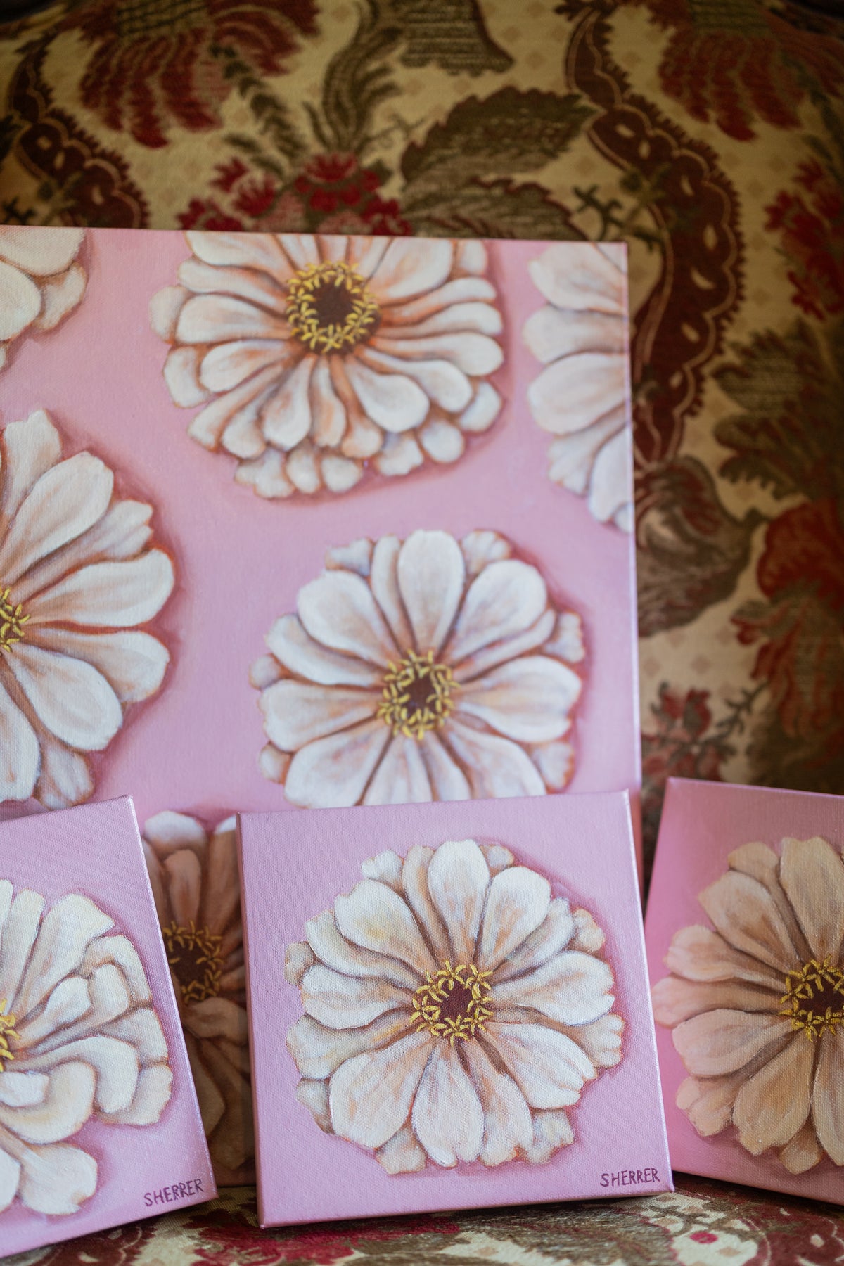 Close-up of white-zinnia paintings on pink, grouped on a patterned upholstered chair.