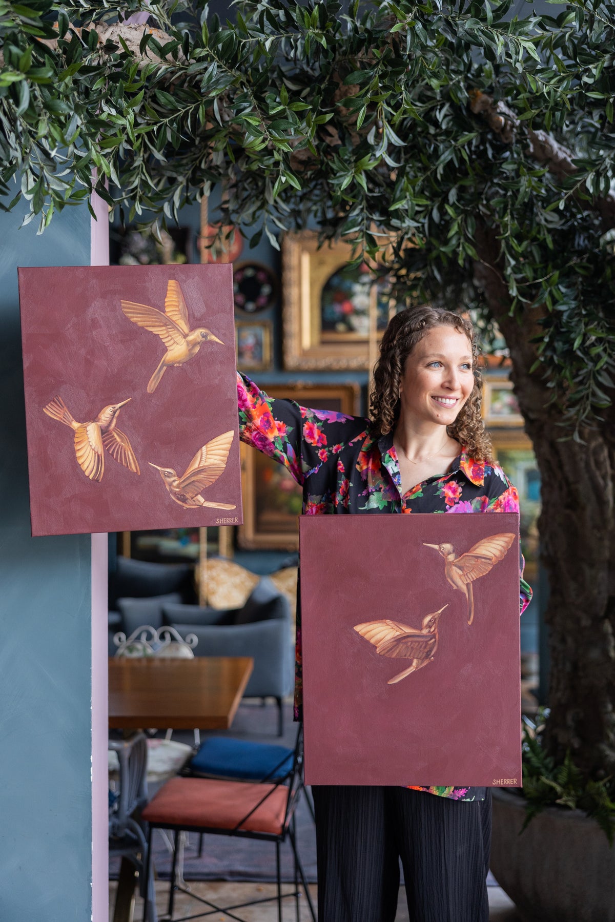 Caroline Sherrer holding two maroon paintings of golden hummingbirds beneath a leafy tree.