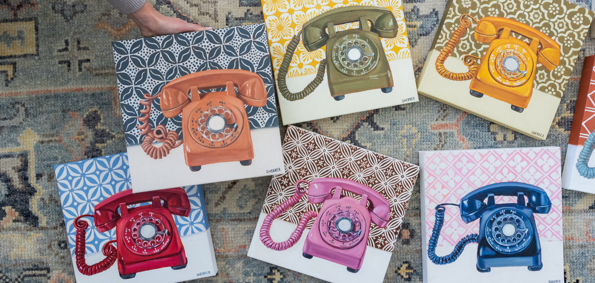 Six square art prints featuring retro rotary phones in different colors, arranged on a patterned rug. A person's hand and arm are visible holding one of the prints, indicating a lifestyle or styled presentation context.
