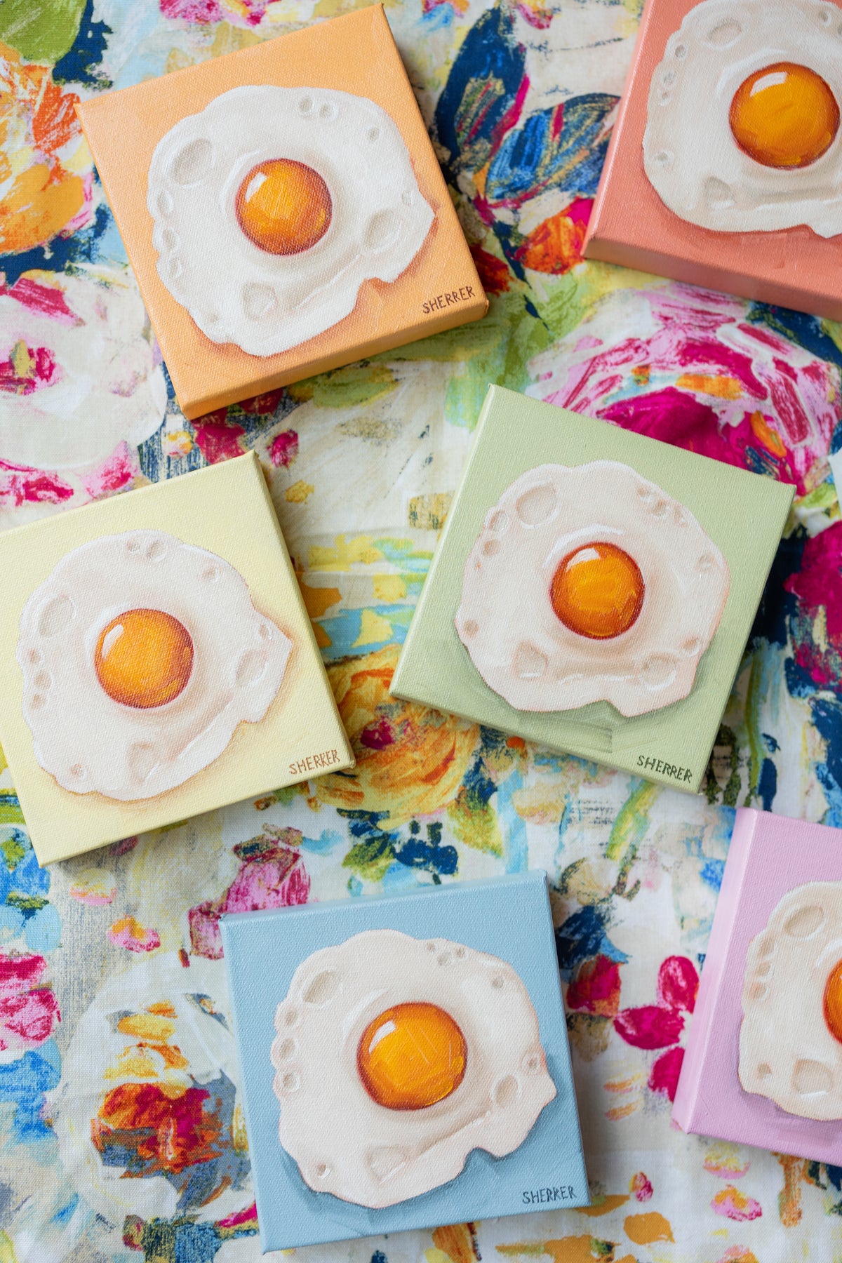 Small fried-egg paintings in yellow, mint, blue and pink arranged on colorful floral fabric.
