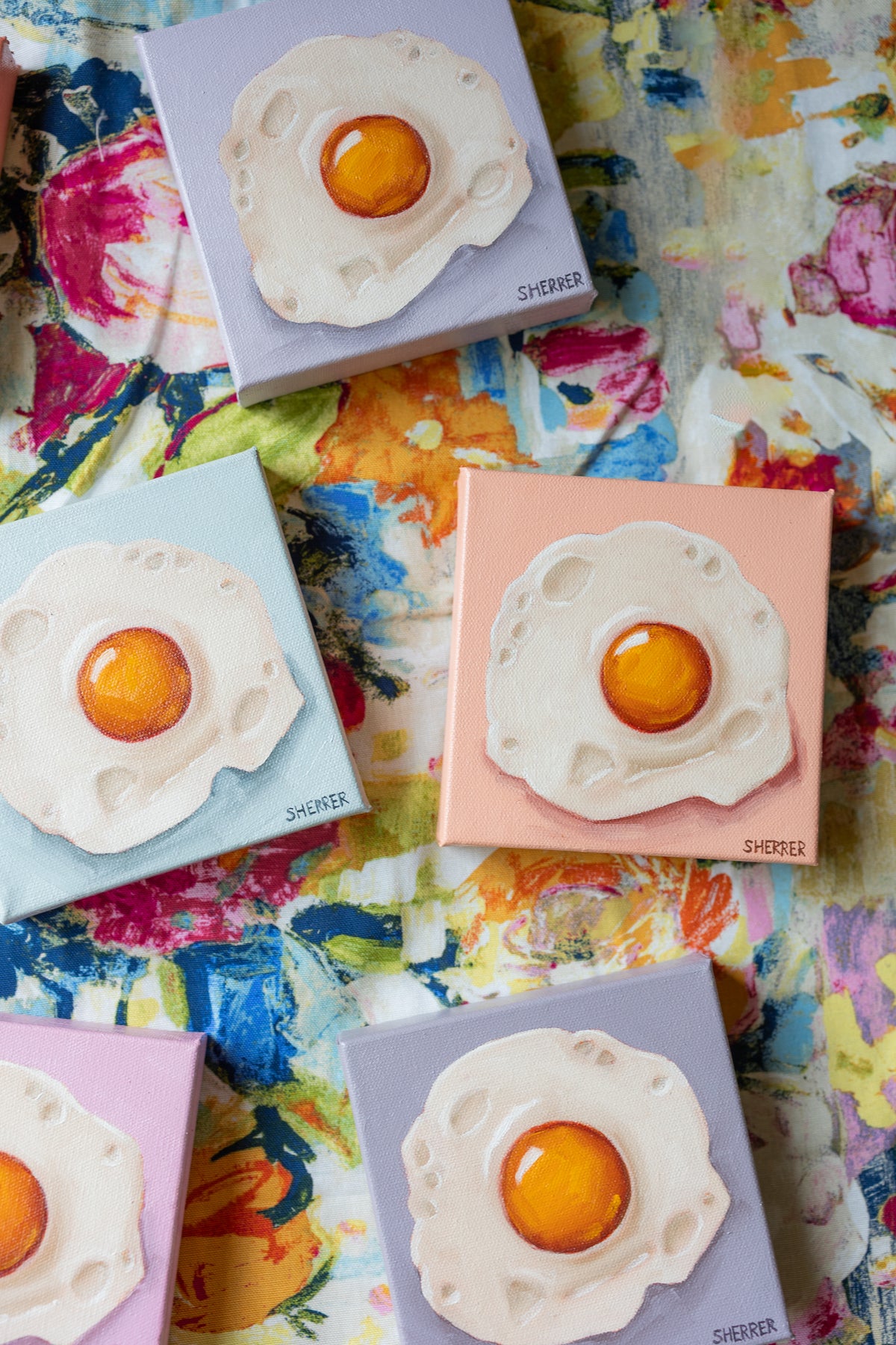 Close-up of small fried-egg paintings on blue, peach and lavender backgrounds arranged on colorful floral fabric.