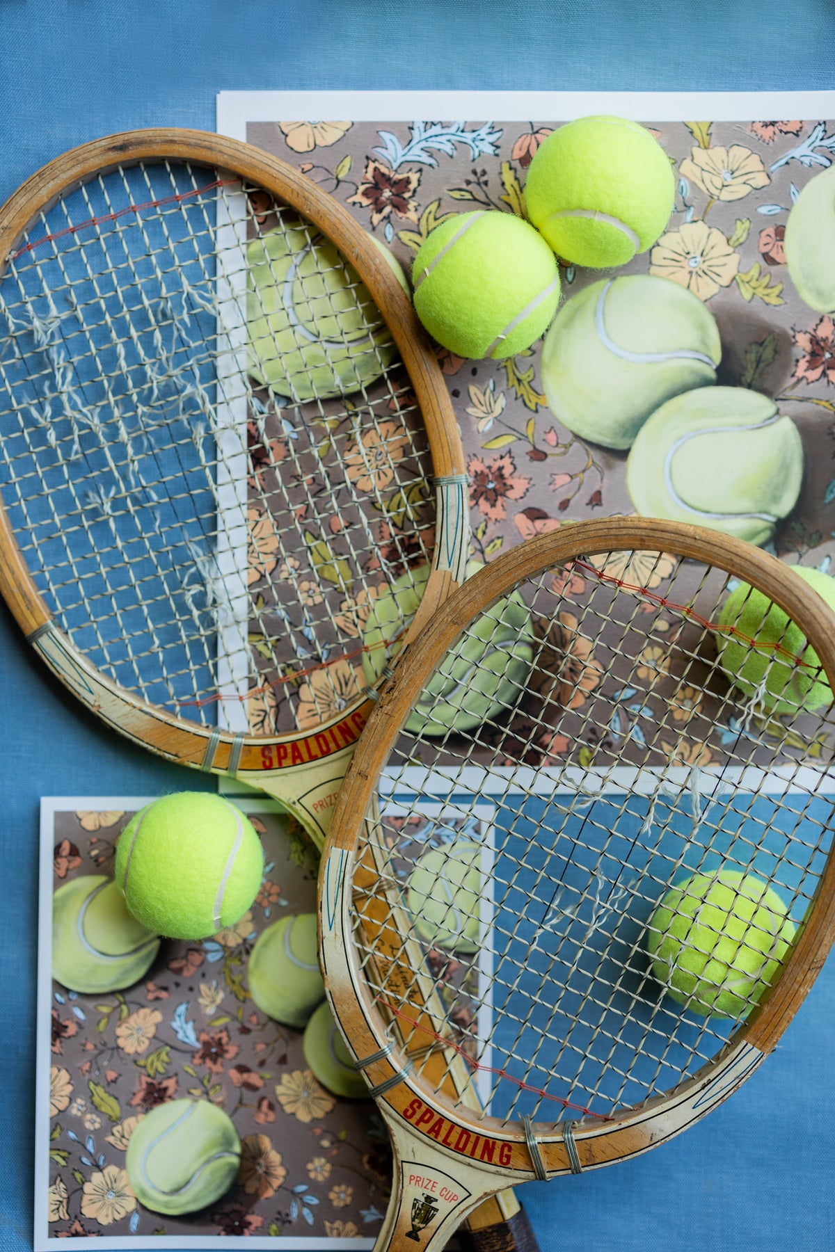 Two wooden tennis rackets and yellow tennis balls layered over white-bordered Weather for Tennis prints on blue fabric.