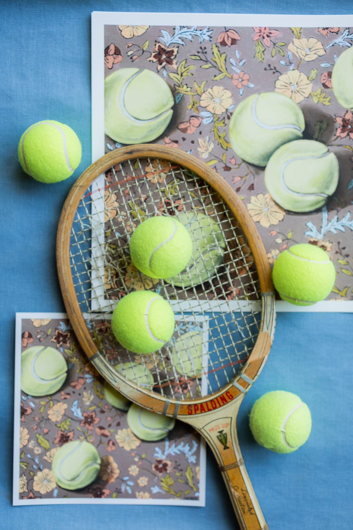 Weather for Tennis prints with white borders arranged on blue fabric beneath a wooden tennis racket and yellow tennis balls.