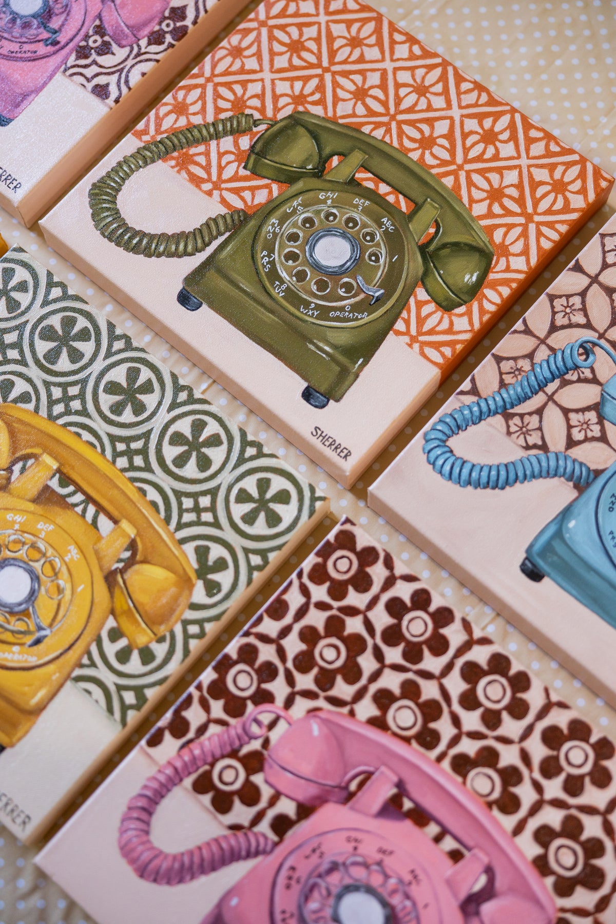 Angled close-up of an olive-green rotary-telephone painting on an orange-and-cream pattern, surrounded by other telephone paintings.
