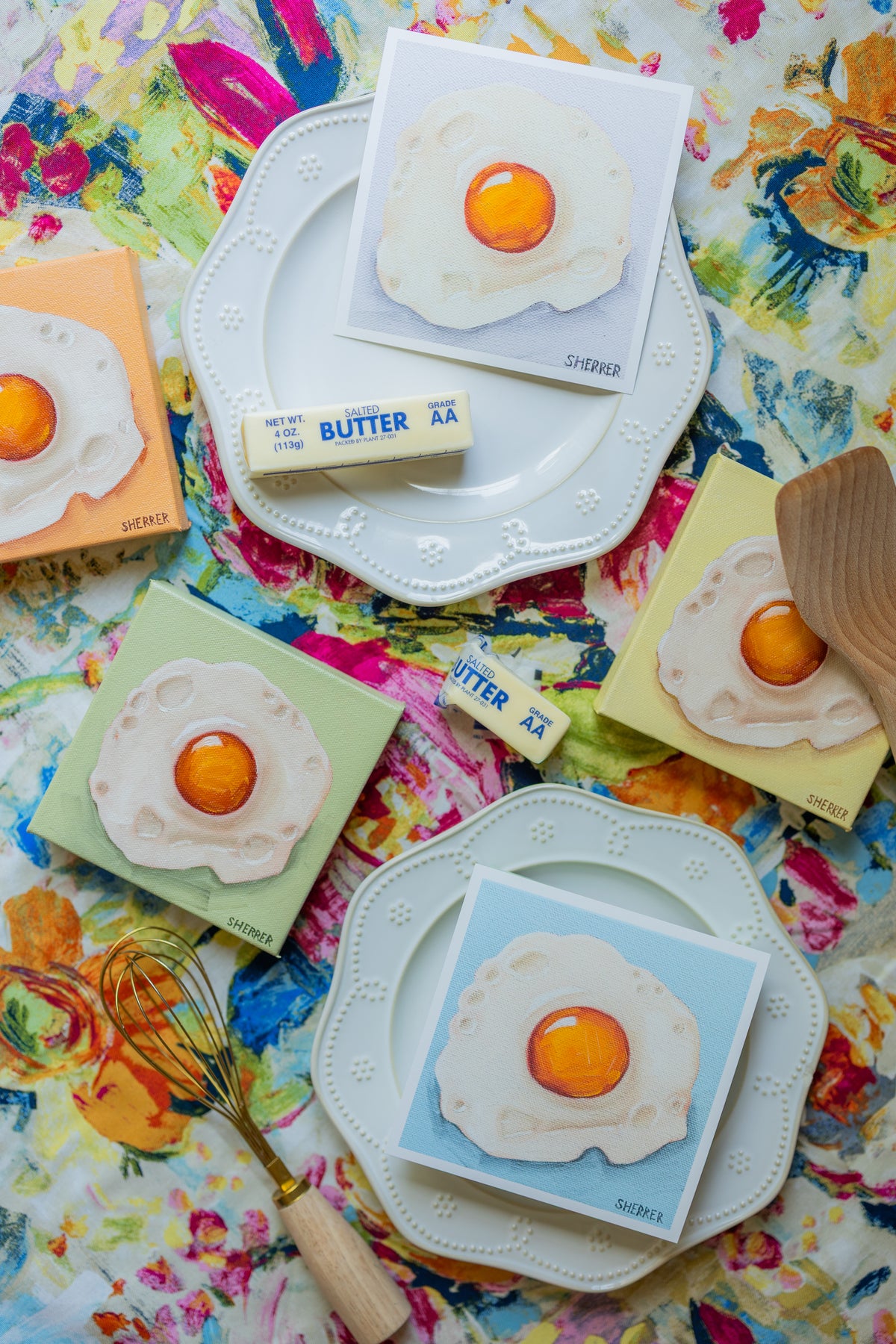 Fried-egg paintings arranged with plates and kitchen utensils on bright floral cloth.