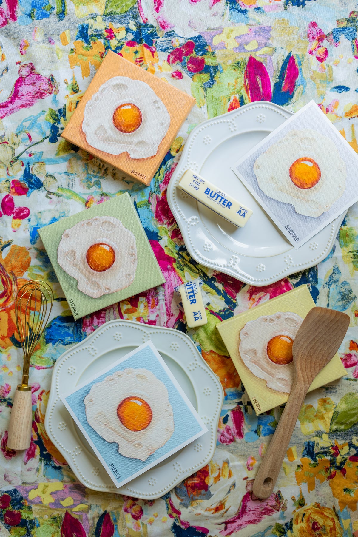 Five small fried-egg artworks in pastel colors arranged with white plates, butter and utensils on colorful floral fabric.