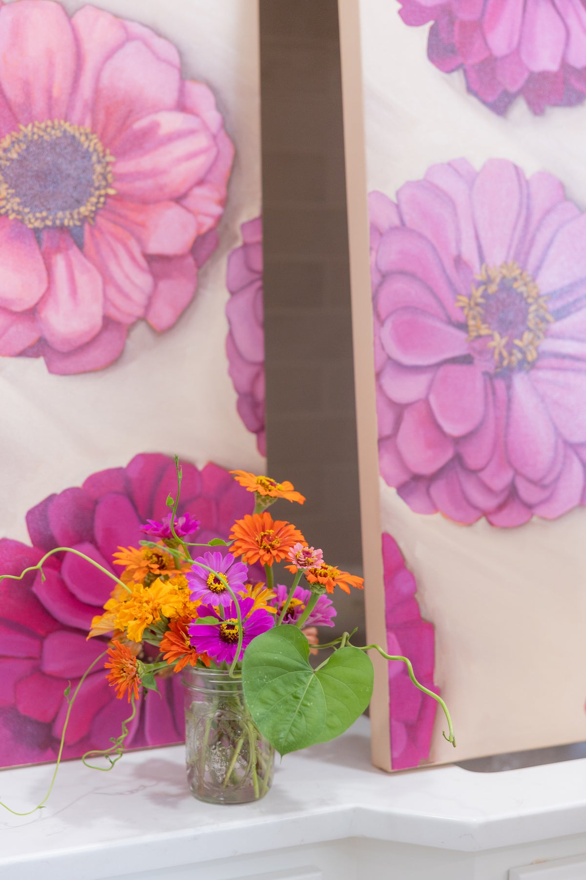 Close-up of pink and purple zinnia paintings standing behind a glass jar of orange and purple flowers.