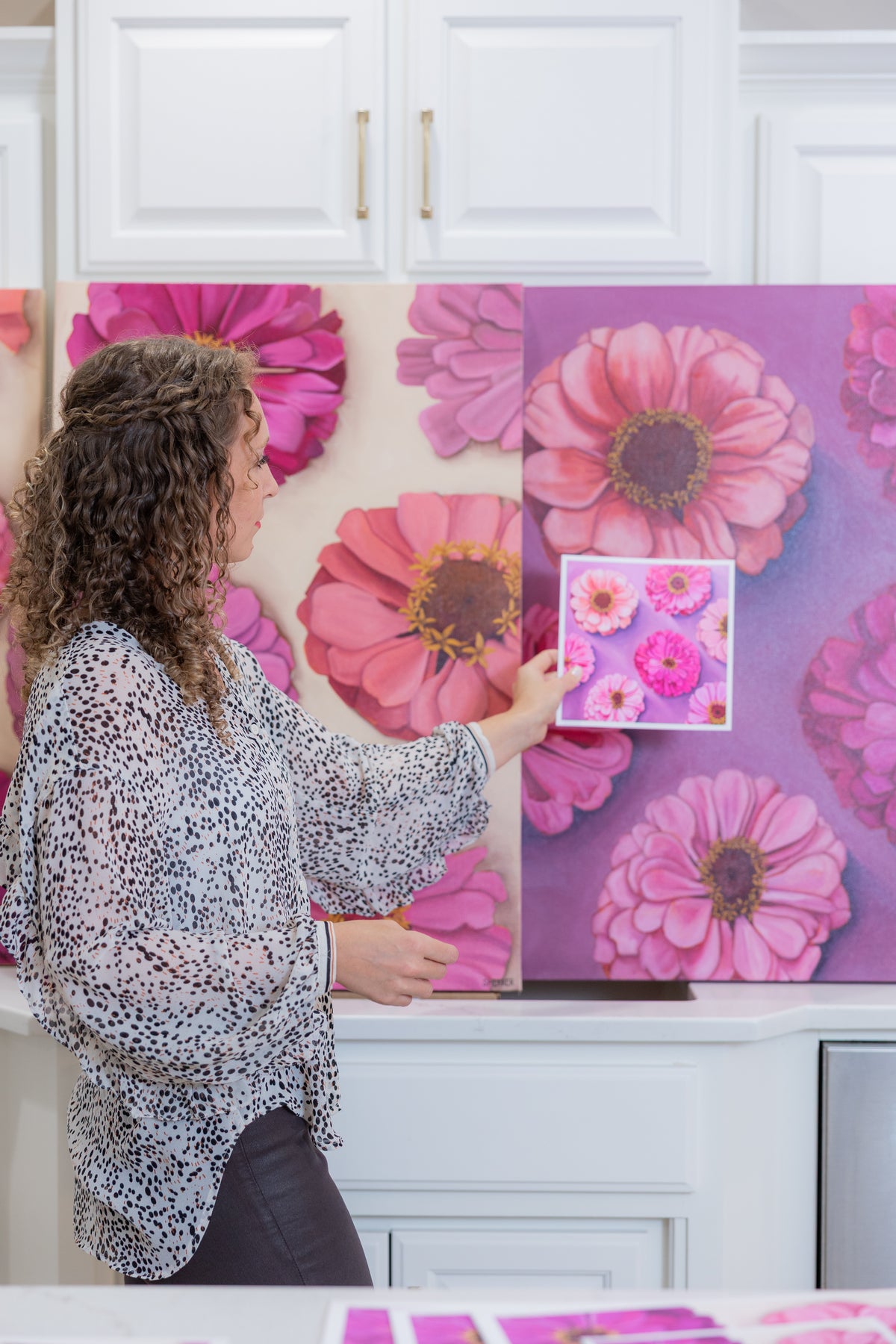 Caroline Sherrer holding a small white-bordered Bold Blooms print in front of the matching large purple-and-pink floral painting.