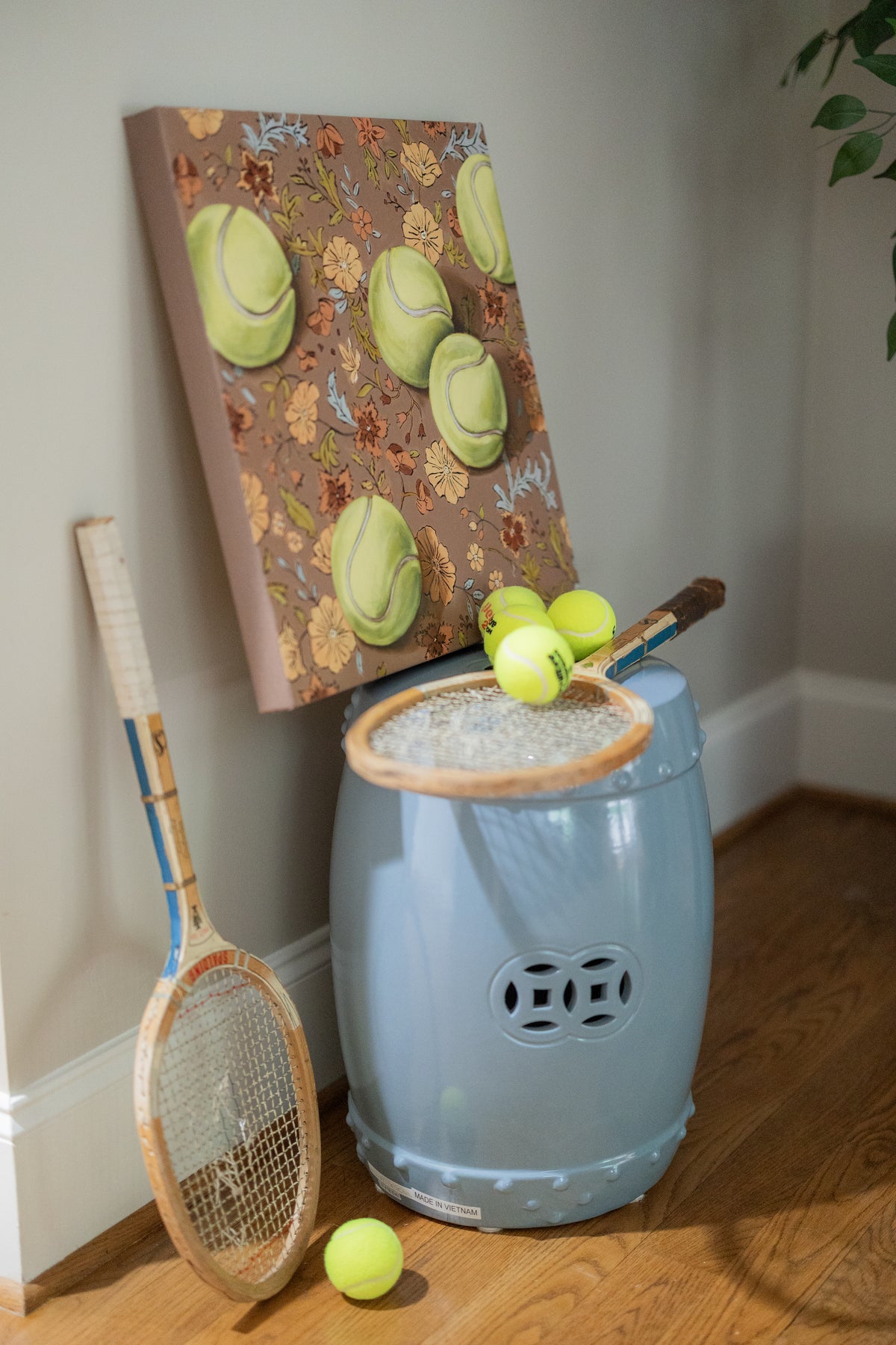 Weather for Tennis painting displayed above a blue ceramic stool with tennis rackets and yellow tennis balls.