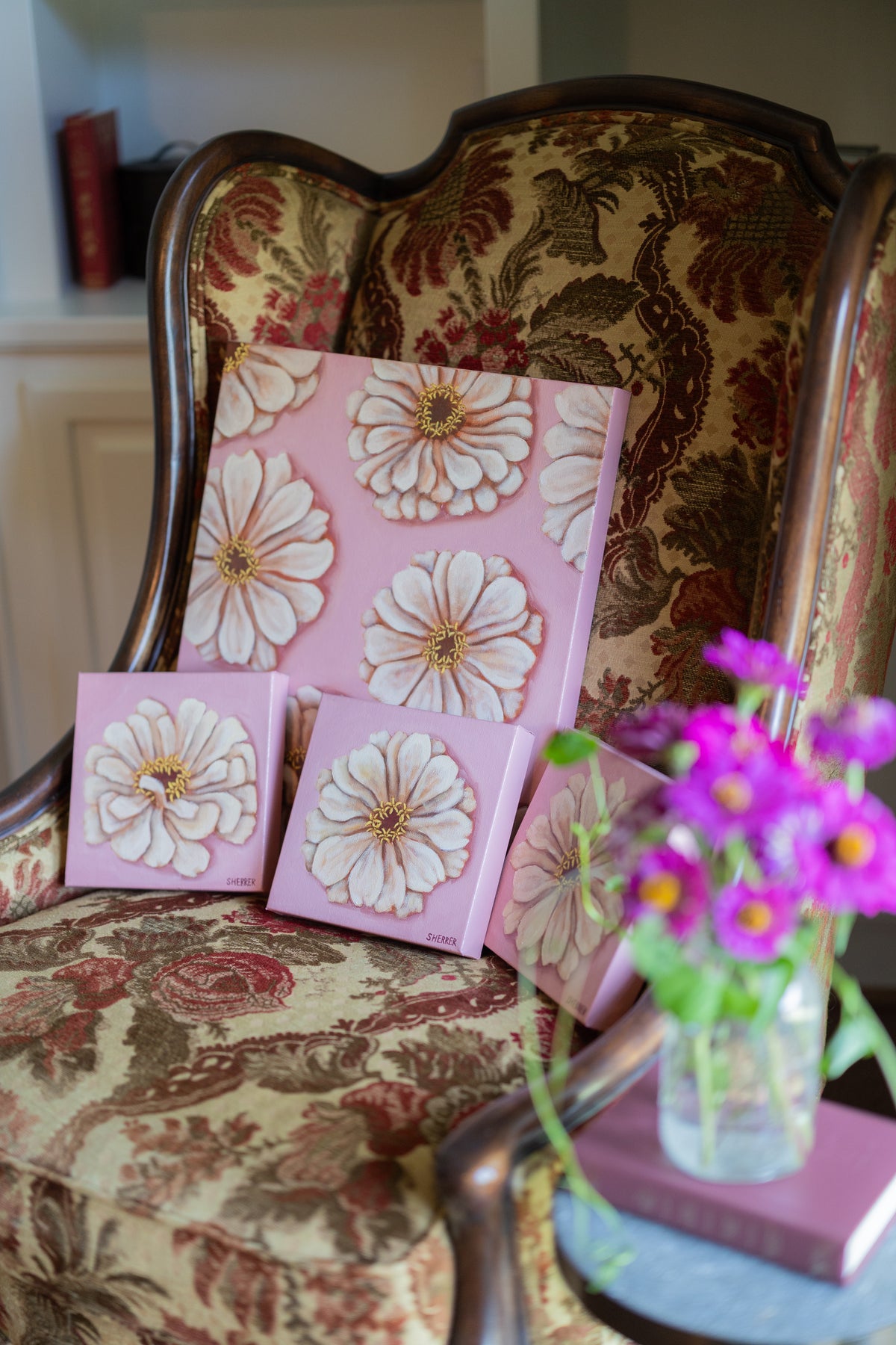 Petal Parade and three small white-zinnia paintings on pink backgrounds arranged on a floral upholstered chair beside fresh flowers.