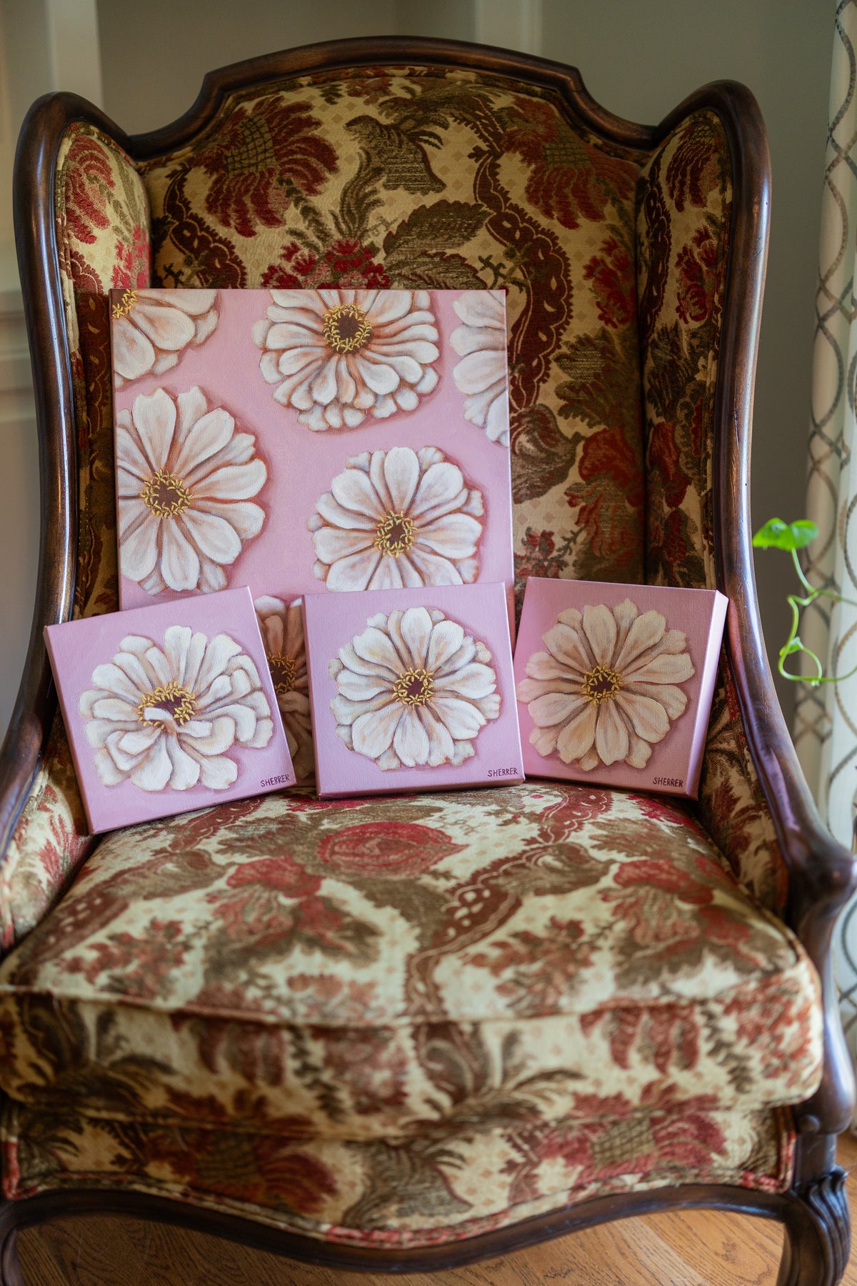 Front view of three small white-zinnia paintings on pink beneath a larger matching floral painting on an upholstered chair.