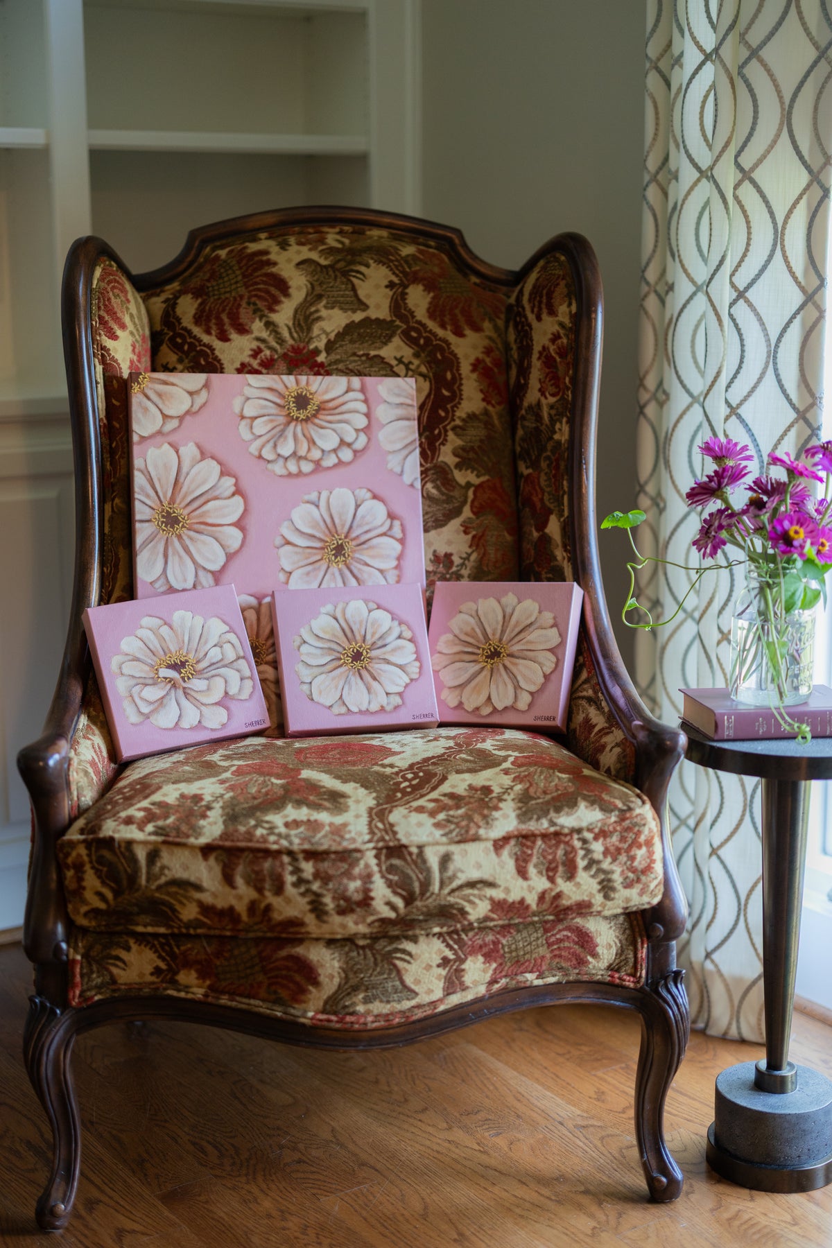 Petal Parade and three small Pink Haze paintings displayed on a floral upholstered armchair beside a vase of purple flowers.