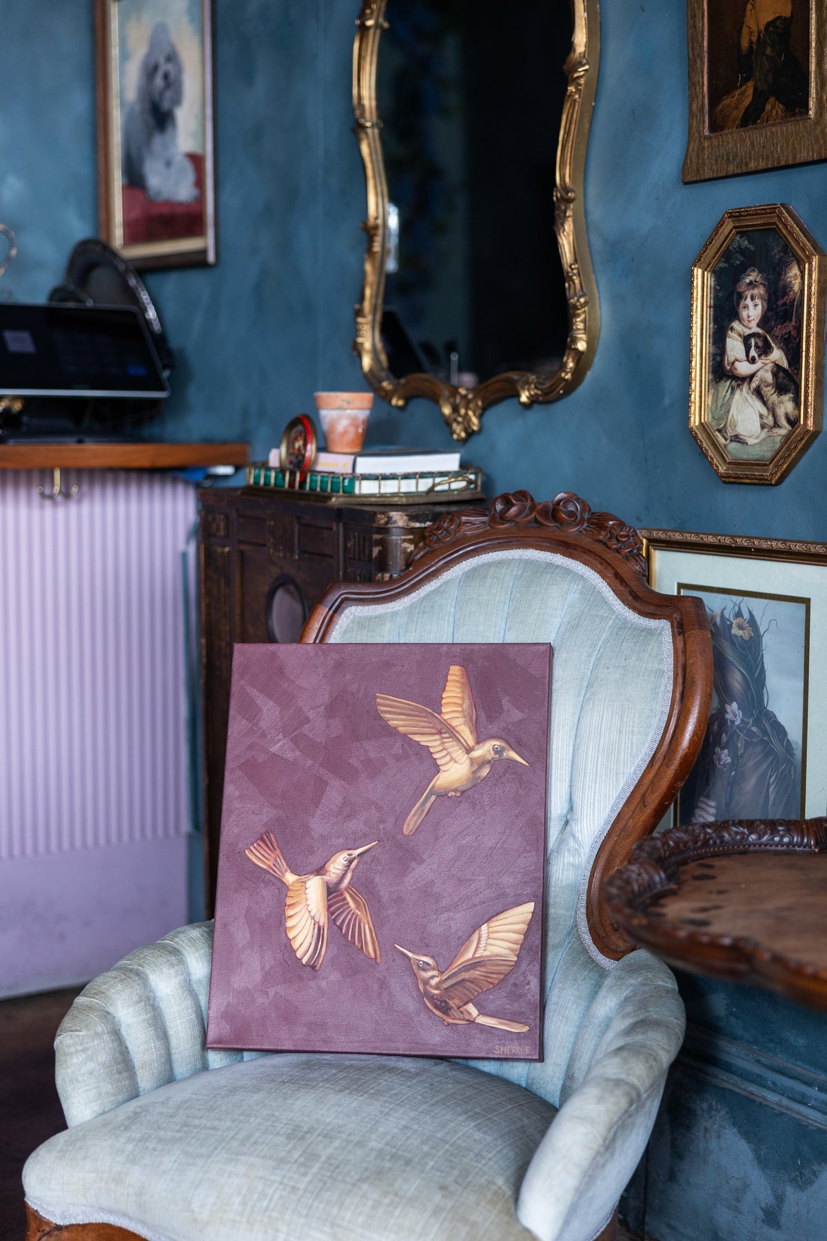 Suspended in Maroon I, showing three golden hummingbirds, resting on a pale upholstered chair in a blue room.