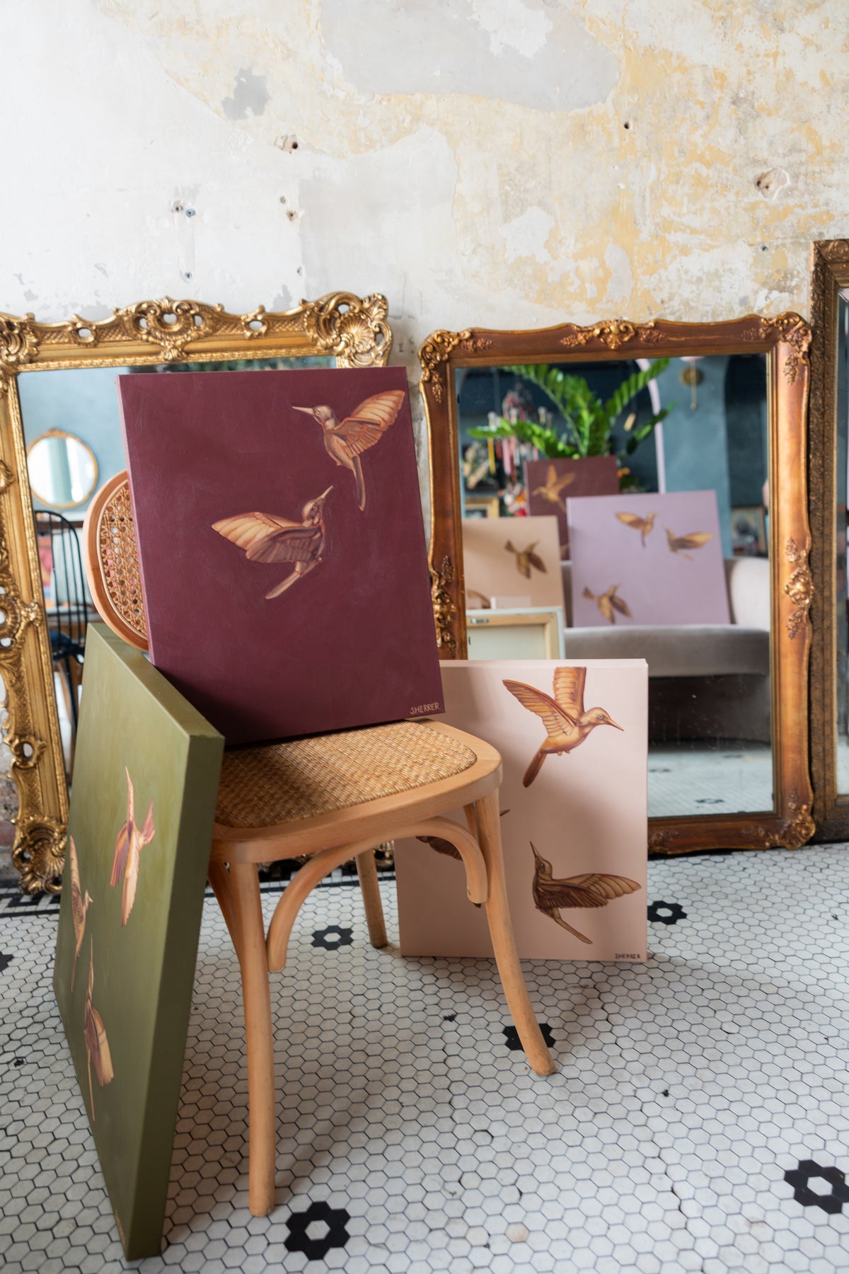 Maroon hummingbird painting resting on a woven-seat chair with other bird paintings in front of ornate gold mirrors.