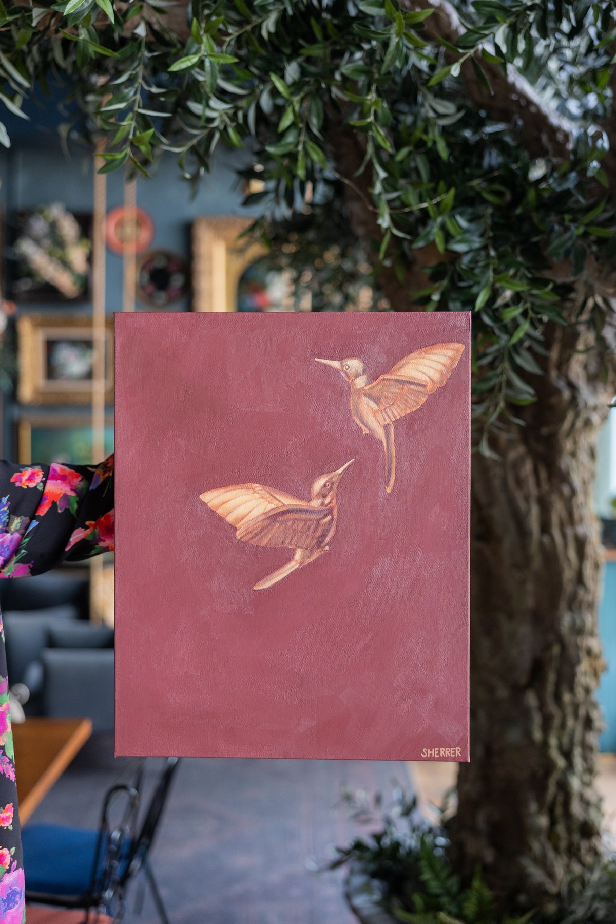 Suspended in Maroon II, showing two golden hummingbirds on maroon, held beside a leafy tree.