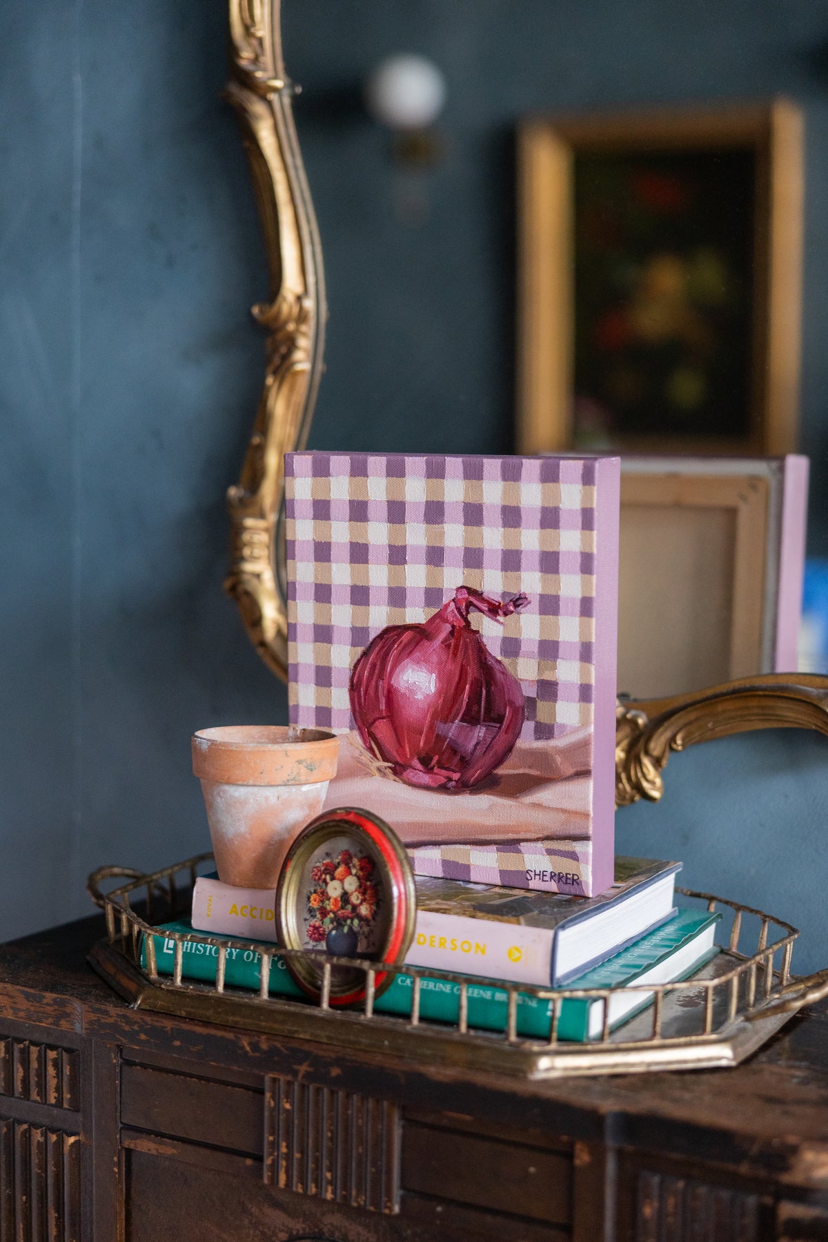 Red Onion on Gingham displayed on stacked books in a brass tray beneath a gold-framed mirror.