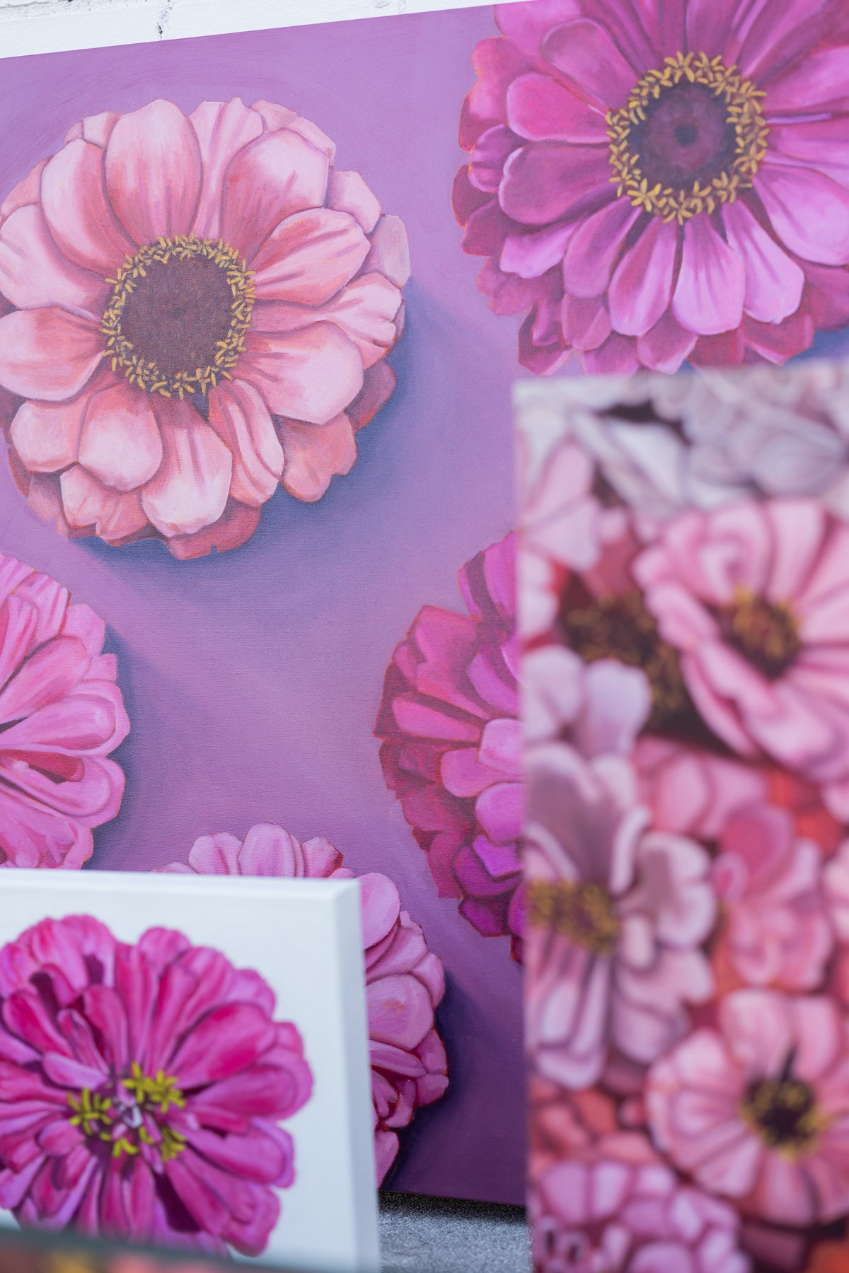Bold Blooms, with large pink zinnias on a purple background, displayed behind other floral paintings.