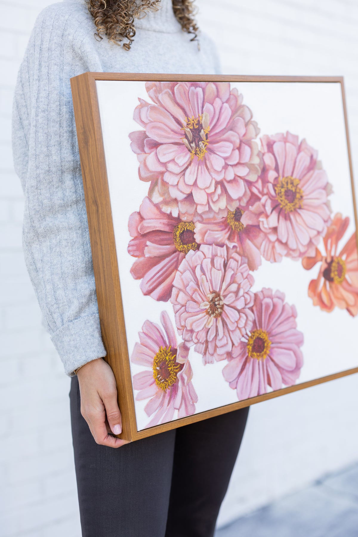 Caroline Sherrer holding the wood-framed Spring Zinnias painting, showing pink and peach flowers on white.